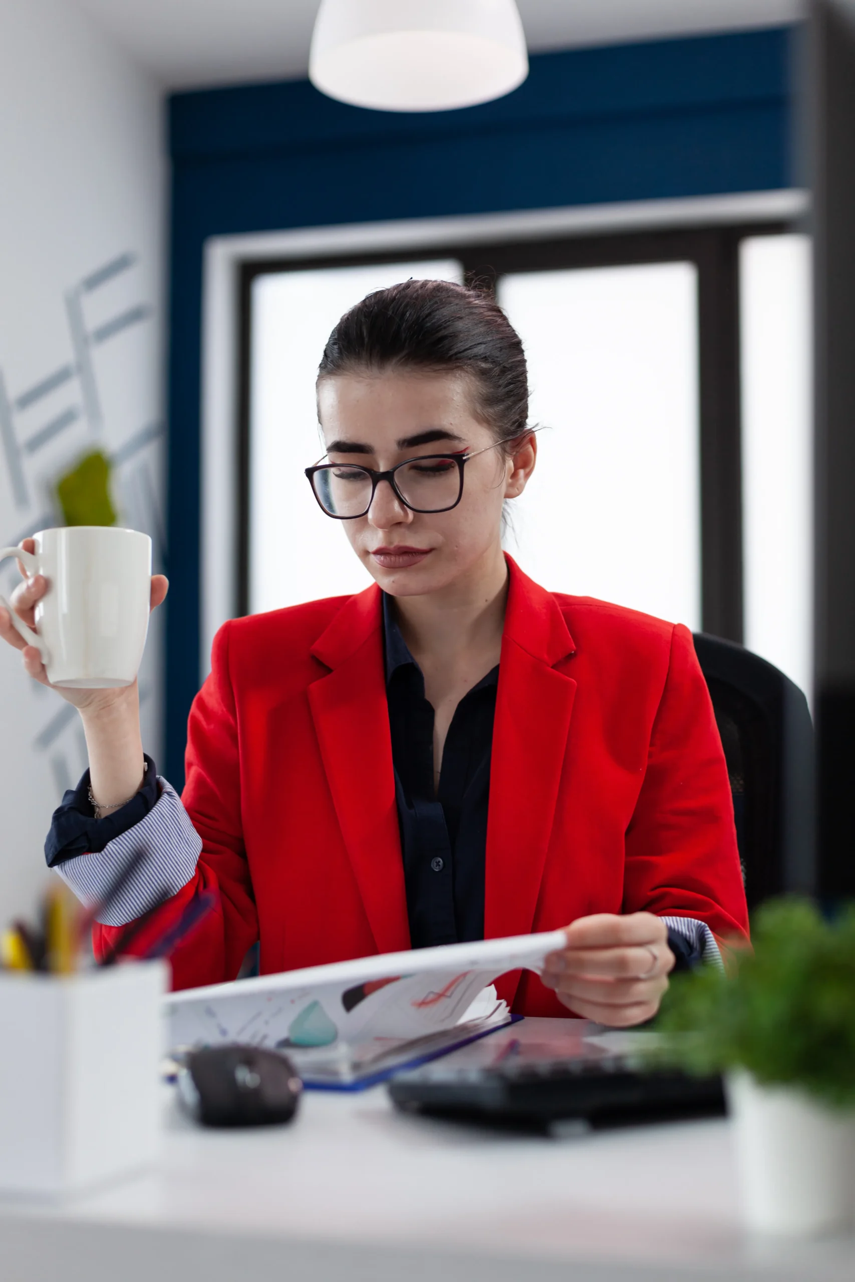 Businesswoman in red blazer reviewing financial documents while holding a coffee cup at her desk, focused on business analysis and planning