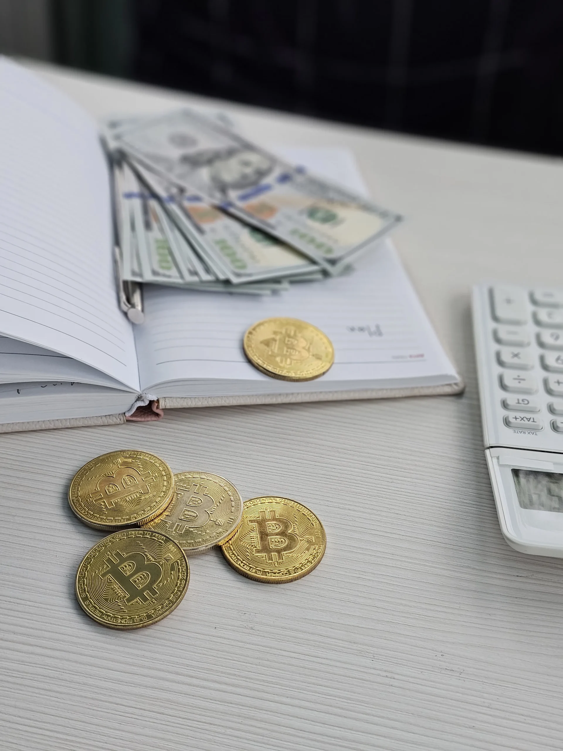 Bitcoin coins on a desk beside a ledger notebook, cash, pen, and calculator, representing crypto taxes, income tracking, and financial planning