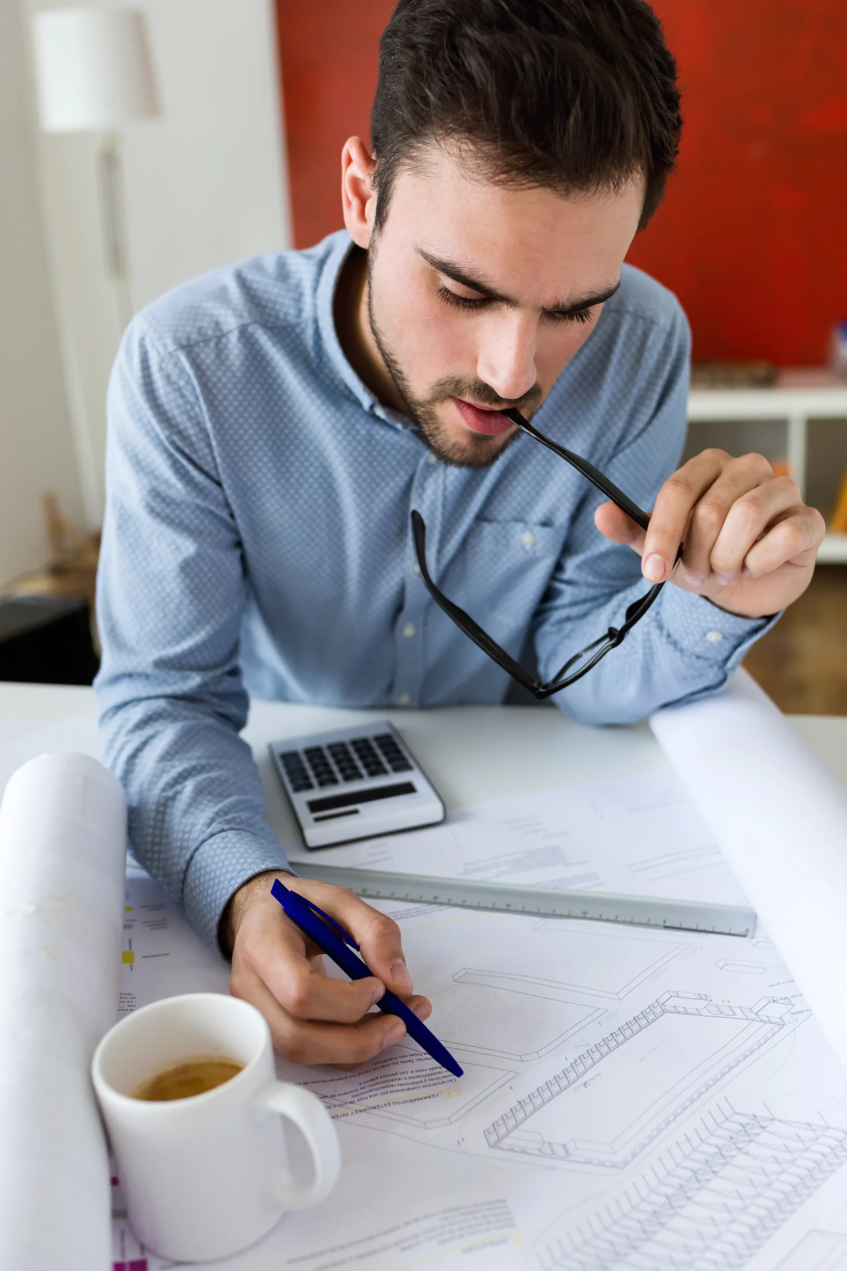 Young professional reviewing architectural plans with calculator and coffee, focused on detailed project analysis at office desk.