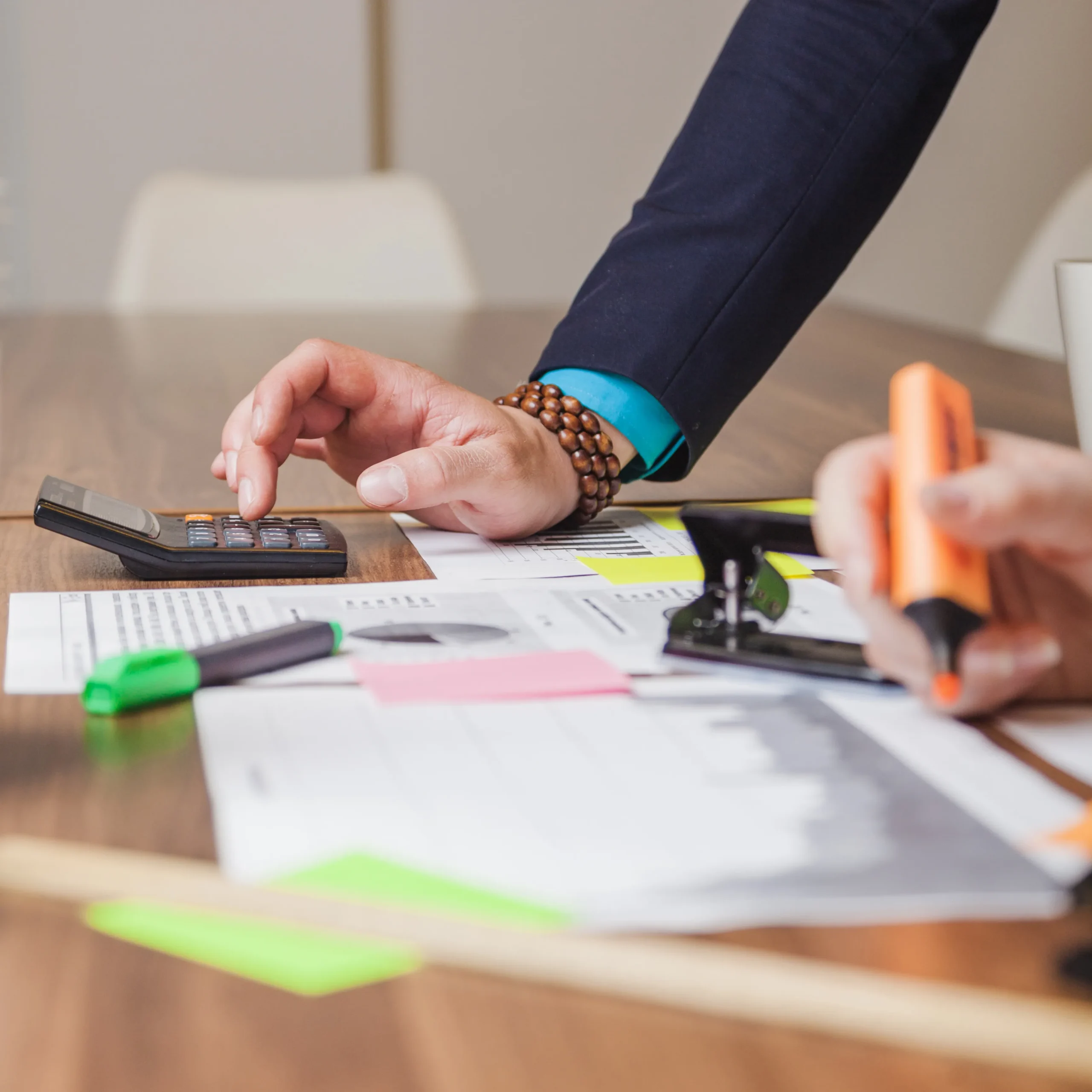 Hands calculating figures on a desk with documents, calculator, highlighters, and stamps, showing focused financial review.